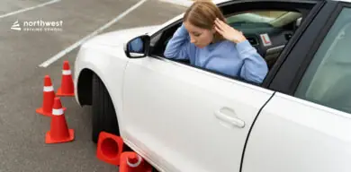 A worried learner driver sits in a white car surrounded by traffic cones, indicating a driving lesson scenario.