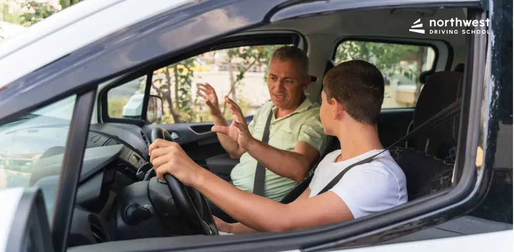 A driving instructor teaches a student in a car, gesturing while the student listens attentively.