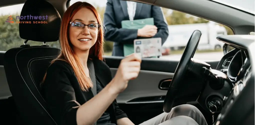 A smiling woman in a car holds up her drivers license while an instructor stands outside the vehicle.