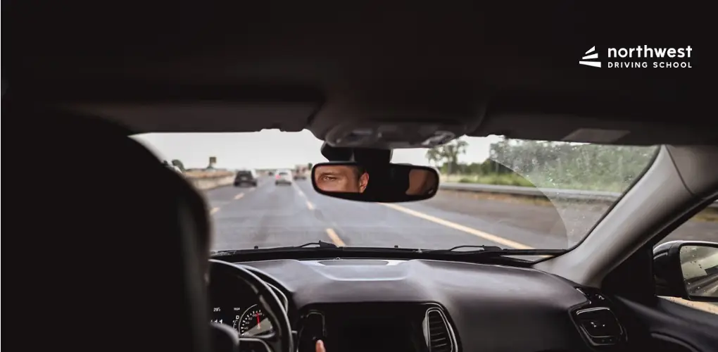 View from inside a car showing a driver focused on the road ahead, with a dashboard and rearview mirror visible.