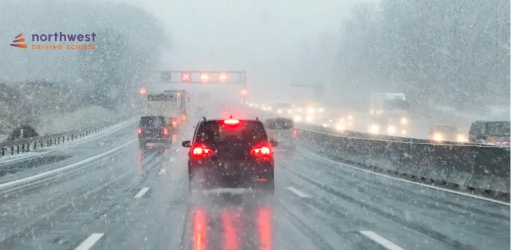 Cars driving on a snowy highway with low visibility and traffic signals ahead.