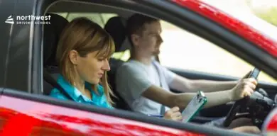 A woman observes and takes notes while a man drives a car during a driving lesson.