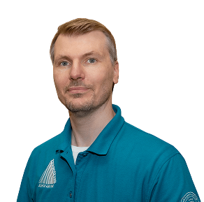 A man with short blond hair wearing a teal polo shirt, looking directly at the camera against a black background.