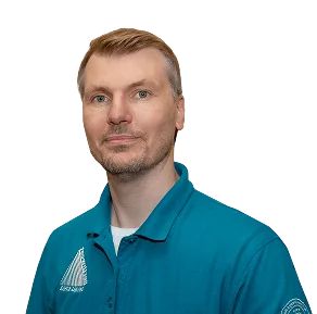 A man with short blond hair wearing a teal polo shirt, looking directly at the camera against a black background.