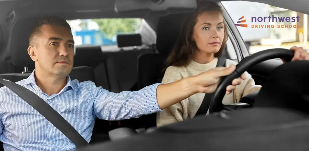 A driving instructor guides a student at the wheel in a car, both focused on the road ahead.