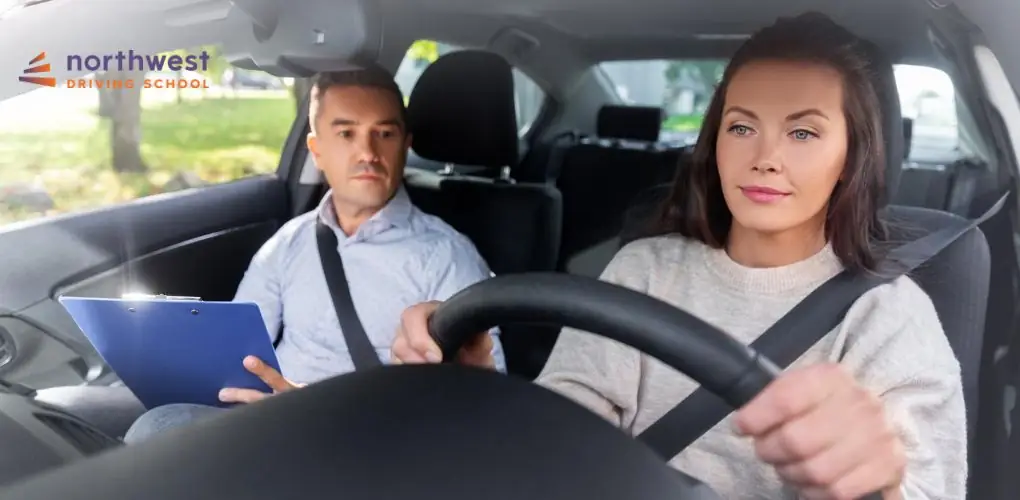 A woman drives a car while an instructor observes and takes notes.