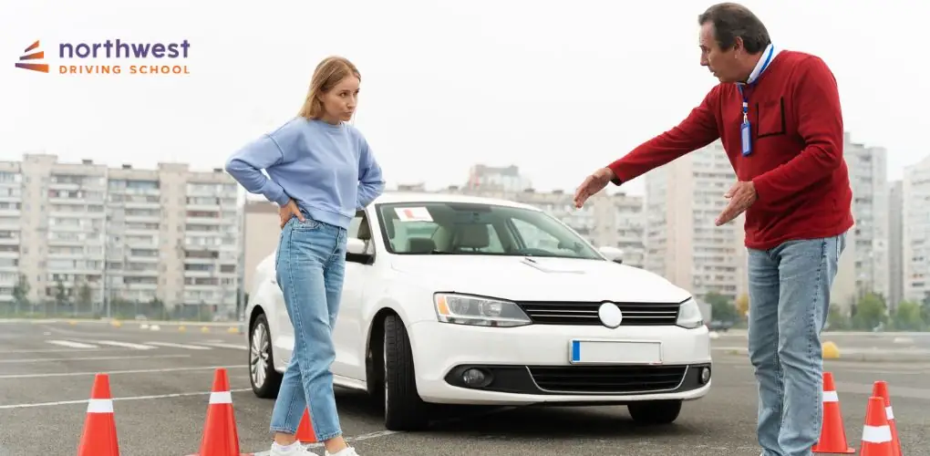 A driving instructor gestures towards a car while a student looks on, surrounded by traffic cones at a driving school.