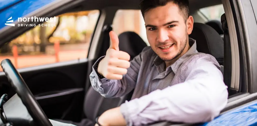 A young man smiles and gives a thumbs-up from the drivers seat of a car, promoting Northwest Driving School.