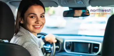 Smiling woman in a car, looking back at the camera, with a driving school logo in the corner.