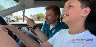 A driving instructor guides a student in a car, pointing while the student smiles and holds the steering wheel.