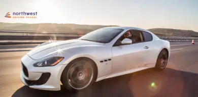 A sleek white sports car drives on a highway with a smiling driver, under a bright sky. Northwest Driving School logo.