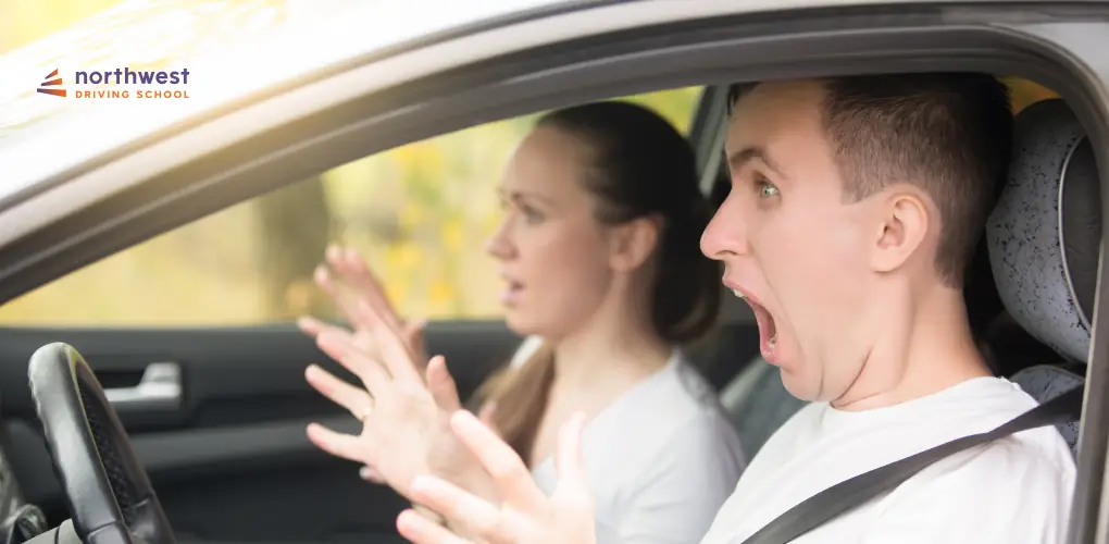 A man in a car looks shocked while a woman appears anxious, both reacting to a driving lesson scenario.