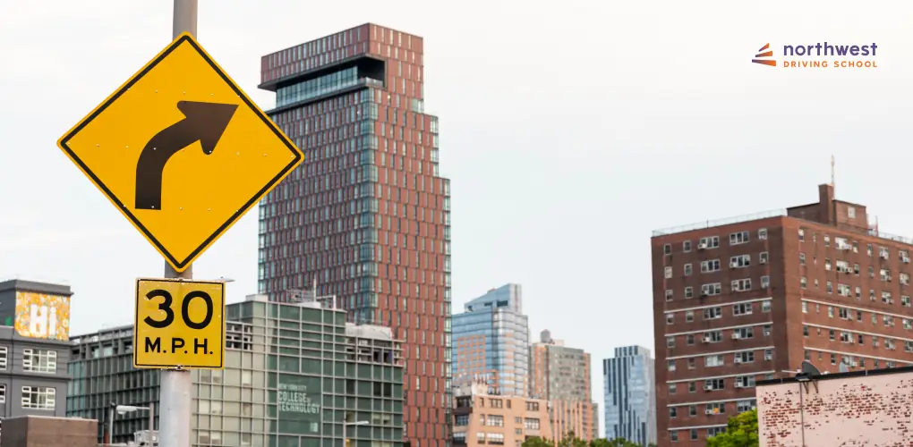 Yellow traffic sign indicating a right turn ahead and a speed limit of 30 MPH, with urban buildings in the background.