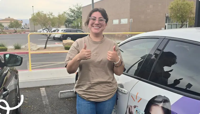 Smiling woman in a beige shirt gives thumbs up beside a parked car in a parking lot.