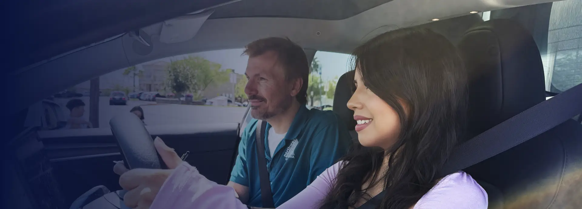 A man and a woman smile while seated in a car, with the woman holding the steering wheel.