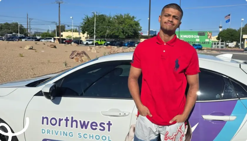 A young man in a red polo stands next to a car with Northwest Driving School signage in a sunny outdoor setting.