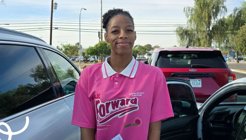 Smiling person in a pink shirt with Walk Forward text, standing beside a car in a sunny outdoor setting.