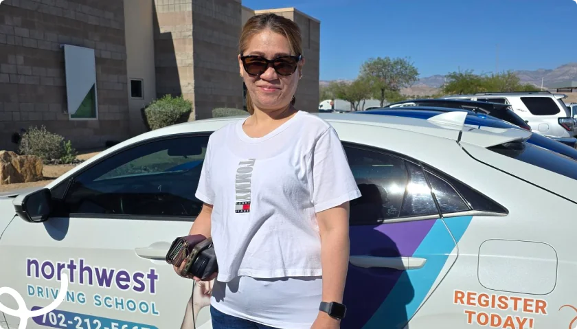 A woman in sunglasses stands next to a car with Northwest Driving School branding in a sunny outdoor setting.